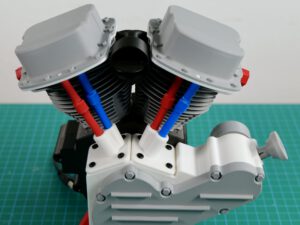 Model two-cylinder engine with dual cylinders and blue/red pushrods, open top on a green cutting mat
