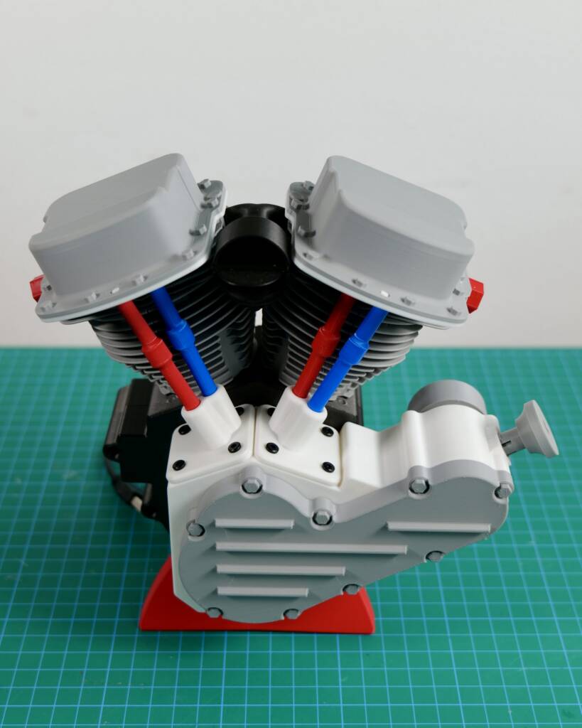 Model two-cylinder engine with dual cylinders and blue/red pushrods, open top on a green cutting mat