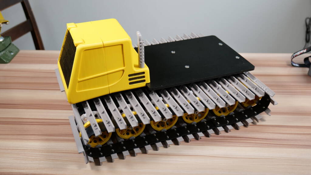 3D-printed RC snowcat track base with yellow cab and black motor platform on a wooden tabletop