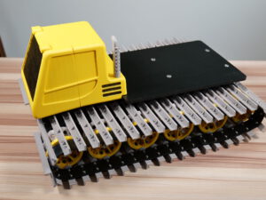 3D-printed RC snowcat track base with yellow cab and black motor platform on a wooden tabletop