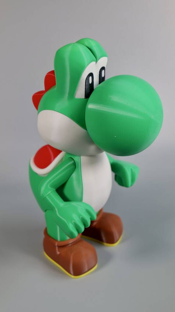 3D-printed Yoshi figurine with green shell and brown shoes posed mid-step against a gray background